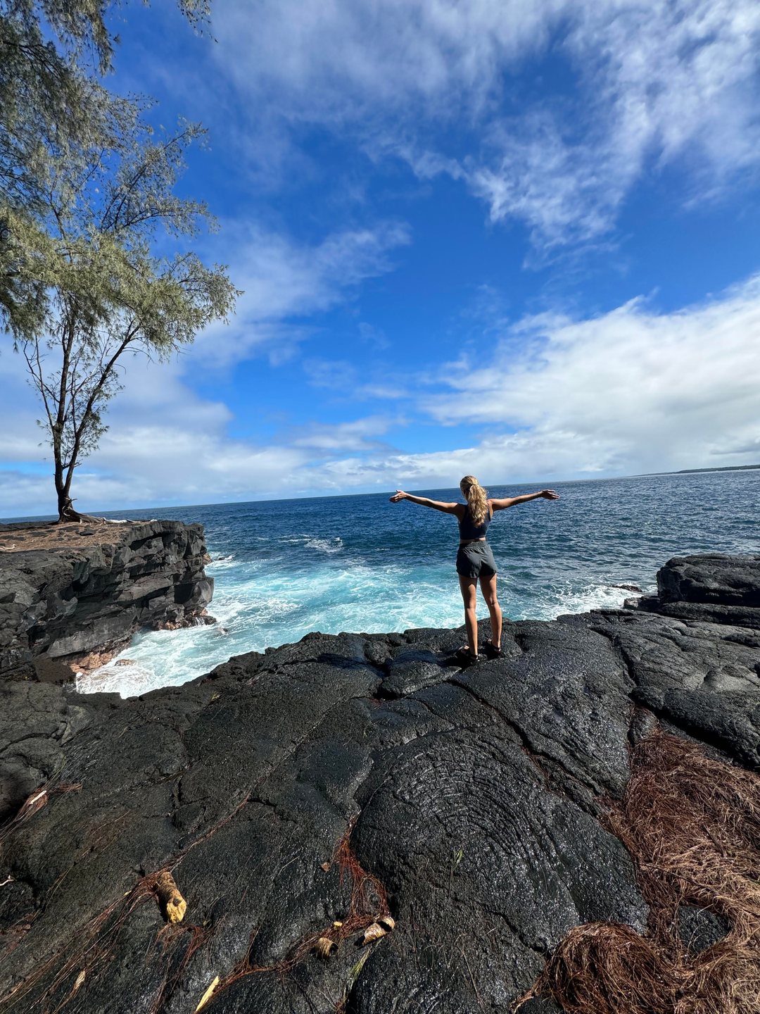 A visitor with arms outstretched on the black lava headland at the Kaloli Point lookout, surf and open Pacific beyond